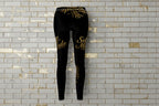 Back view of Self Made gold script black leggings on a grey brick background, showcasing women's compression activewear.