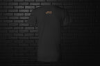 Back view of a black Unbothered Vibe Women's V-Neck Graphic Tee with gold logo on a dark brick background.