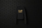 Black CocoMe Couture fleece hoodie with gold dripping logo on a dark brick background — luxe streetwear front view.