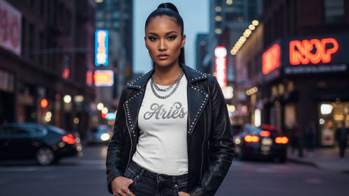 Model in streetwear wearing white Aries zodiac sign tee with leather jacket and silver chains on a city street at night.