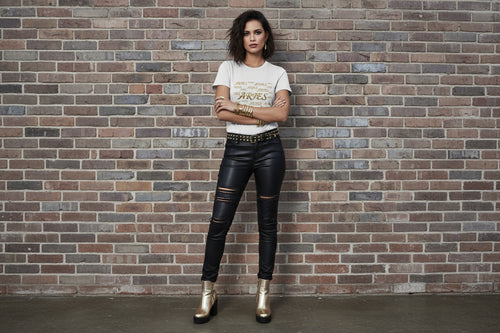 Woman in Aries the Ram Premium Tee with gold zodiac graphic and black moto pants against a brick wall background.
