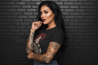 Tattooed woman wearing a Soft but Dangerous Performance Tee activewear top against a black brick wall.