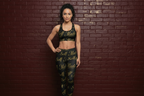 Woman in Self Made gold script black leggings and sports bra set standing against a dark brick wall.