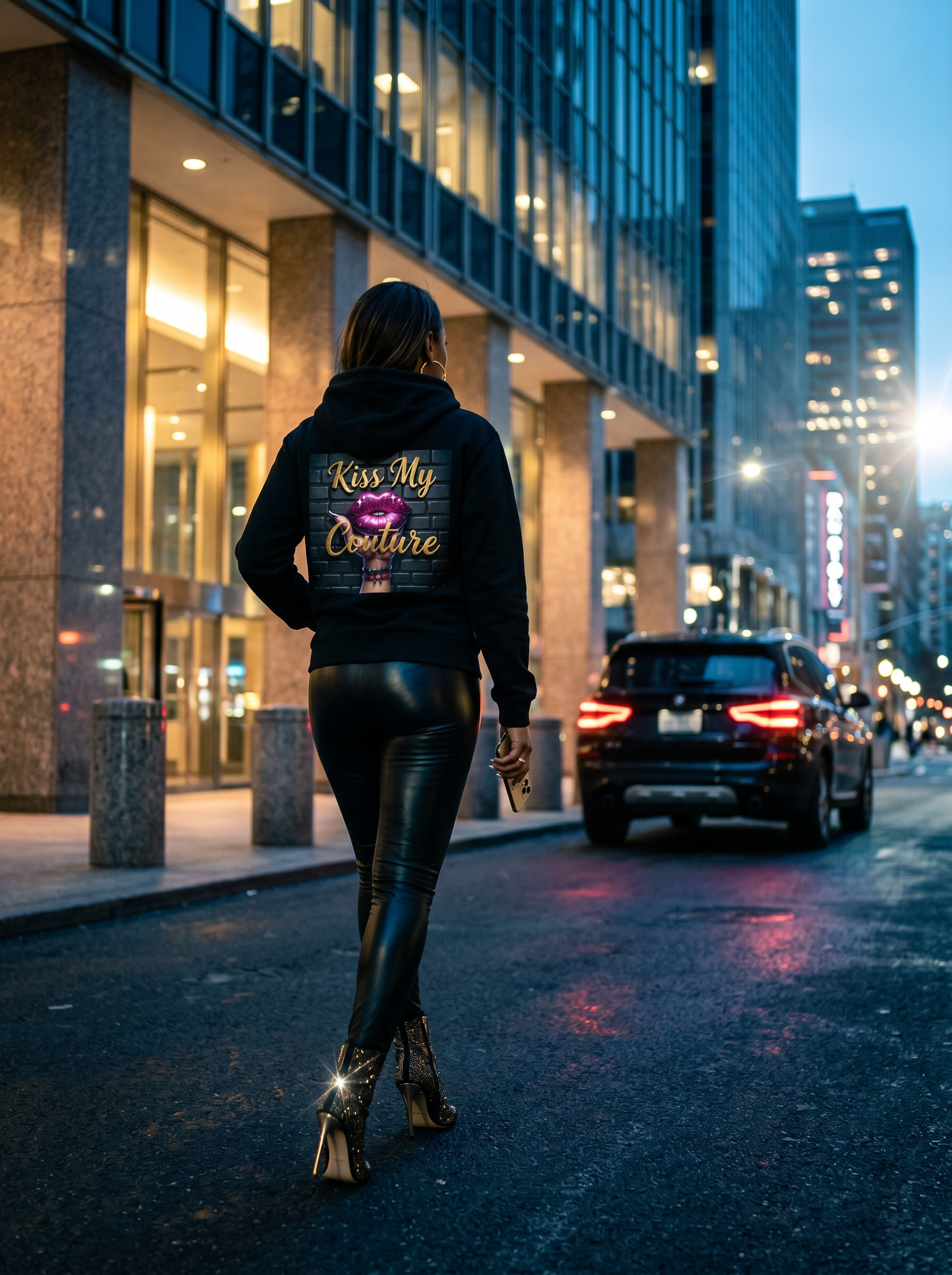 Woman in Kiss My Couture Rebel-Luxe Fleece Hoodie and leather pants, walking away on a city street at dusk.